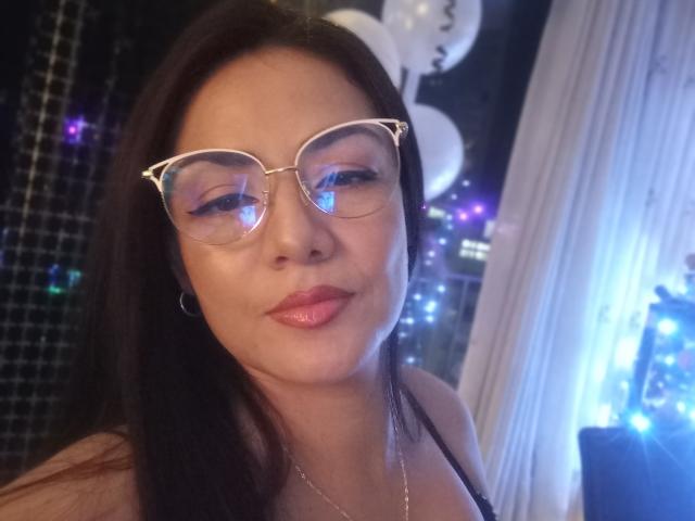 KendraHeat, usando óculos, com lábios rosados e sensuais, pele brilhante, cabelo preto, vestindo uma blusa preta, pequenos brincos de argola e um colar de prata, em um fundo com iluminação suave..