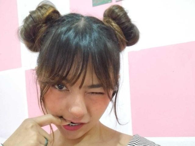 PaulinaaSweet has brown hair in two buns, with straight bangs covering her forehead. She's winking and biting her finger. Her skin is light, and she has pink lipstick. She wears a striped top. The background is a pink and white checkerboard pattern. Her hair has a glossy, smooth texture, and her expression is playful. The image is a photograph.