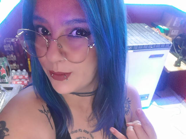 DarkLilith with blue hair, glasses, nose ring, and lip piercings, showing tattoos on her shoulders and chest, in a selfie, with a small fridge in the background.