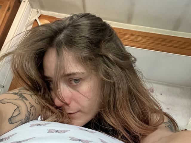 LexireyLoverEver with messy brown hair, green eyes, tattoos on arm, lying down, looking up, wearing a white butterfly-patterned top, slightly biting bottom lip.
