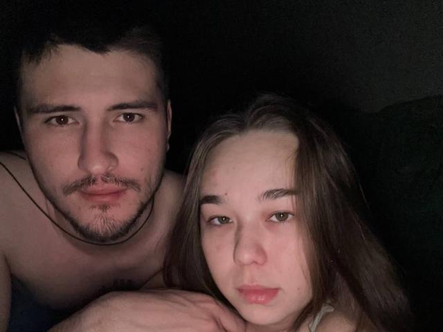 ZaxodisYuda, shirtless with a beard, has a serious face. ZaxodisYuda, with long brown hair and pouty lips, leans against him. Both have pale skin and dark eyes.