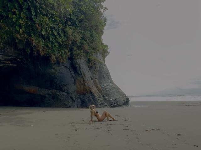 VictoriiaGrey, naked, blonde, small tits, sitting on sandy beach with rock cliff and green foliage on left, overcast sky, wet sand, small waves in background.