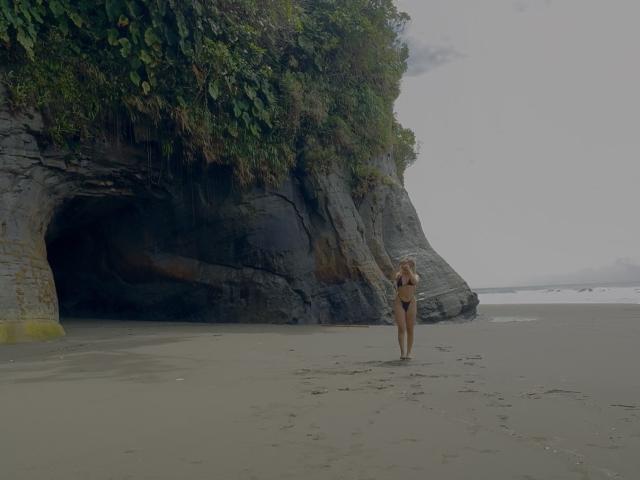 VictoriiaGrey stands alone on a sandy beach, wearing a tiny black bikini, with her ass and small tits visible, near a large rocky cave with green foliage.
