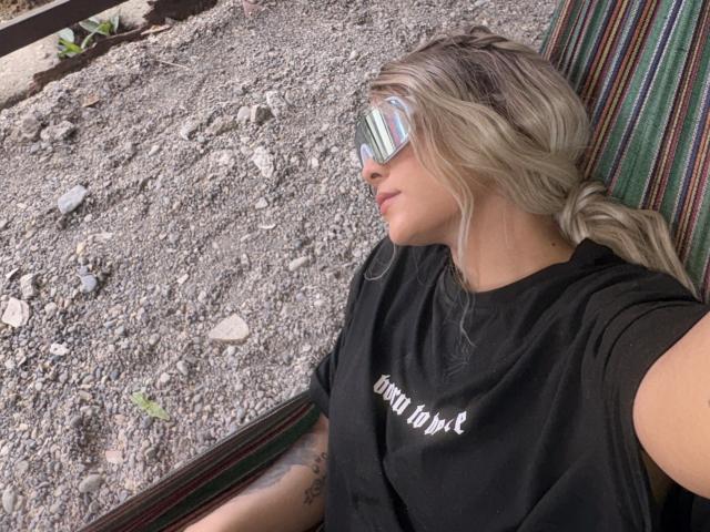 VictoriiaGrey with blonde hair, wearing reflective sunglasses, black shirt, and striped hammock, lying on gravel with small rocks, right side profile, relaxed, tattoo on arm.