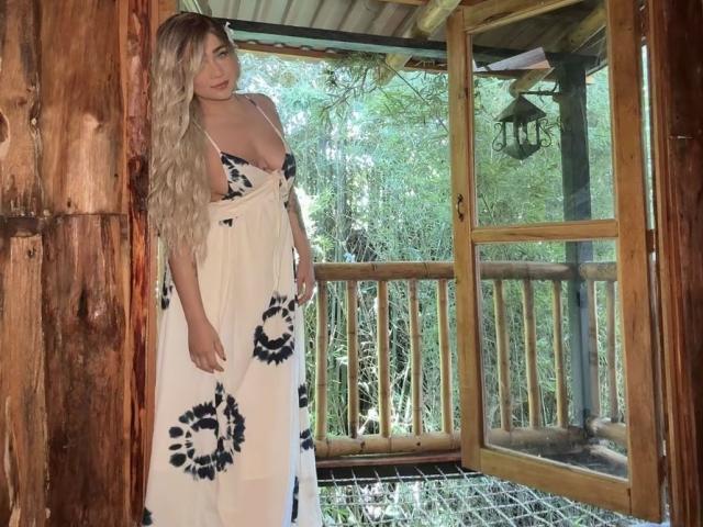 VictoriiaGrey stands in a wooden doorway, wearing a white dress with black floral patterns, exposing her big tits. She has long blonde hair and a cute face.