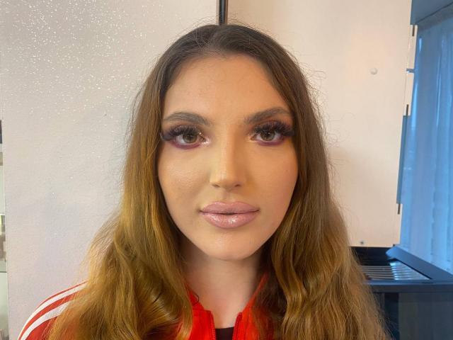 LaraSynn has long brown wavy hair, pink glossy lips, and thick, long, dark eyelashes. She's wearing a red and white striped shirt, looking straight at the camera.
