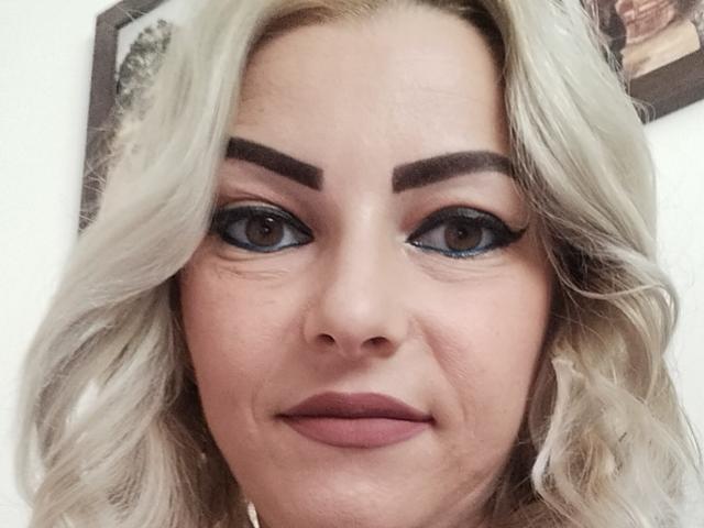 KatyDirty tem cabelo loiro ondulado, sobrancelhas escuras, delineador azul e batom rosa-marrom. Seus olhos castanhos olham diretamente para a câmera com um sorriso discreto..