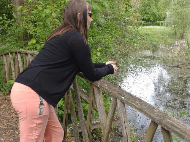 YviEngel87-hot leans on a wooden bridge, wearing a black sweater and pink pants with a butt plug keychain. She has brown hair and is looking at a water-filled area.