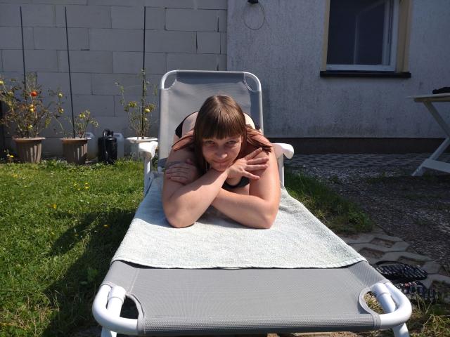 YviEngel87-hot, nude, on a gray lounge chair with arms folded under chin, on a white towel, lying on grass, brown hair, small tits, flirty expression, sunny backyard.