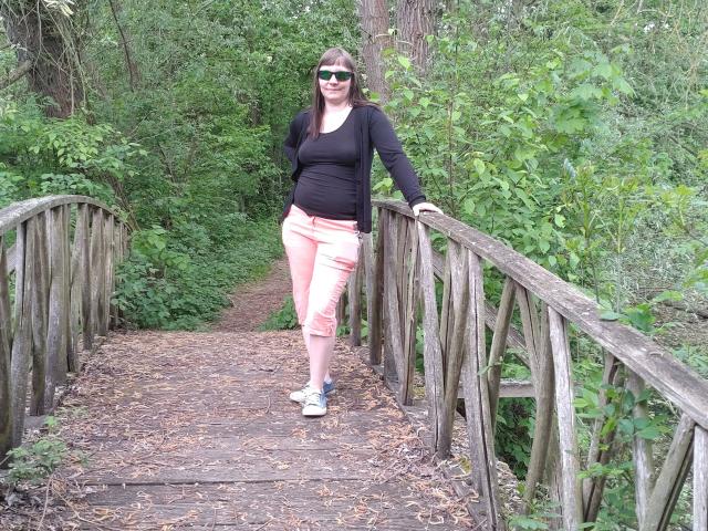 YviEngel87-hot stands on a wooden bridge, wearing a black top, pink pants, white sneakers, and green sunglasses, with a hand on the railing, surrounded by greenery.