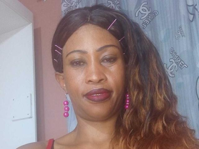 NaughtyCakex has medium brown skin, wavy brown hair with pink hairpins, pink earrings, and dark purple lipstick. She's wearing a red top, looking slightly to the left.