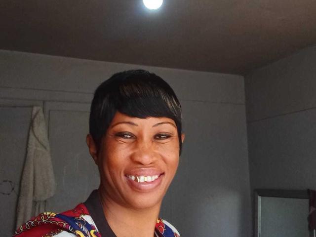 NaughtyCakex with short black hair, smiling with white teeth, wearing a colorful patterned shirt, in a room with a ceiling light.