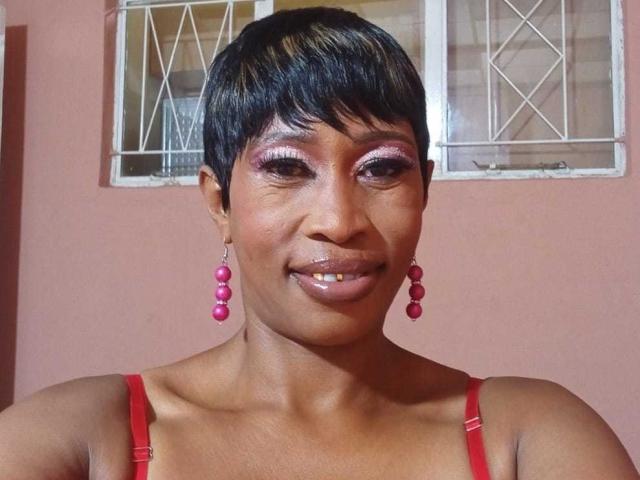 NaughtyCakex has short black hair, shiny pink eye makeup, glossy lips, and wears red earrings and a red strap top. She's smiling with a cute, confident look.