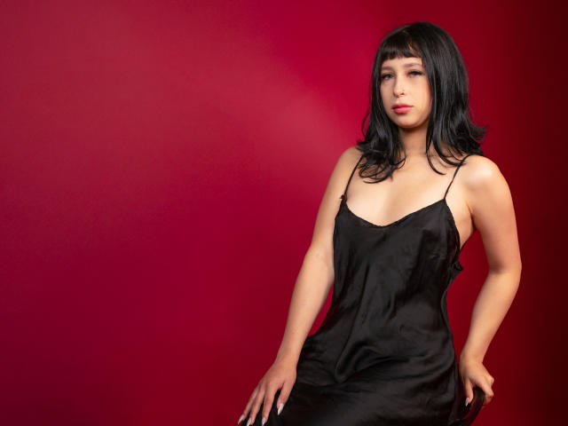 Dalia, con i capelli neri e ondulati, indossa un abito nero di seta che mette in mostra il seno, seduta di fronte a uno sfondo rosso, guarda leggermente di lato con un'espressione neutra..