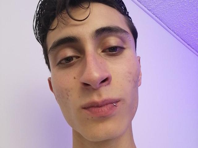 IthanSkin ha un viso pieno di brufoli, sopracciglia scure, capelli neri corti e umidi, e un labbro inferiore con un piercing. Lo sfondo è di un colore viola chiaro. La pelle è pallida con piccole macchie di acne..