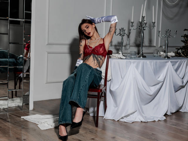 SophieKitty met tatoeages, rode kantbeha, witte handschoenen, groene jeans, zwarte hakken, zittend op een stoel, armen omhoog, tafel met kaarsen en een witte doek op de achtergrond..