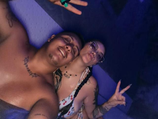 JackAndTriana are topless, Jack has tattoos and a smile, Triana has glasses, tattoos, and a peace sign, both making a goofy face, Jack has a medium-sized, flaccid penis.