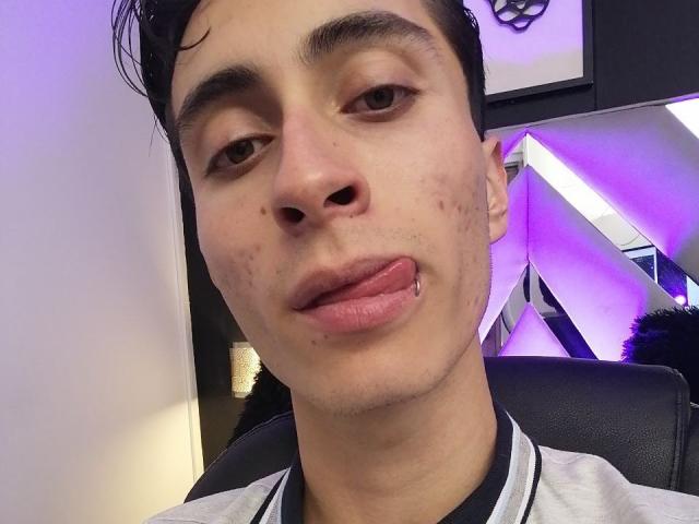IthanSkin a une peau claire, des cheveux courts et foncés, et un piercing à la lèvre où sa langue est visible. Il porte une chemise grise et noire..