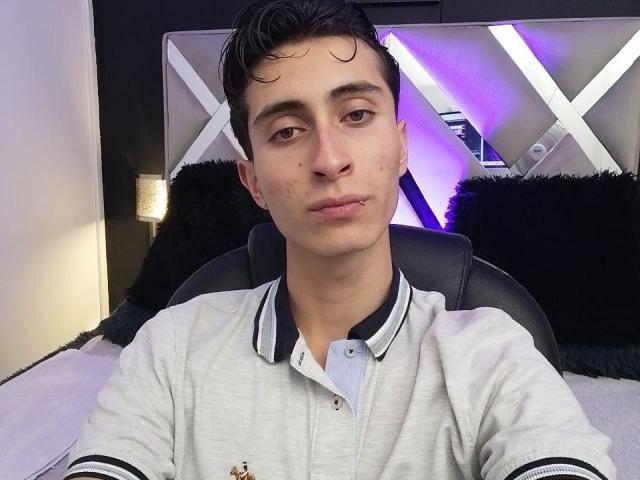 IthanSkin ha un leggero sorriso, capelli neri corti e una polo grigia con bottoni e bordi neri. È seduto su una sedia nera e ha un piccolo piercing al labbro..
