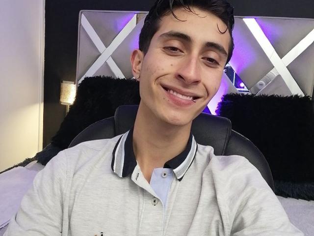 IthanSkin with short dark hair, smiling, wearing a gray polo shirt, black collar, and black ear piercing, sitting on a black leather chair, in a well-lit room.