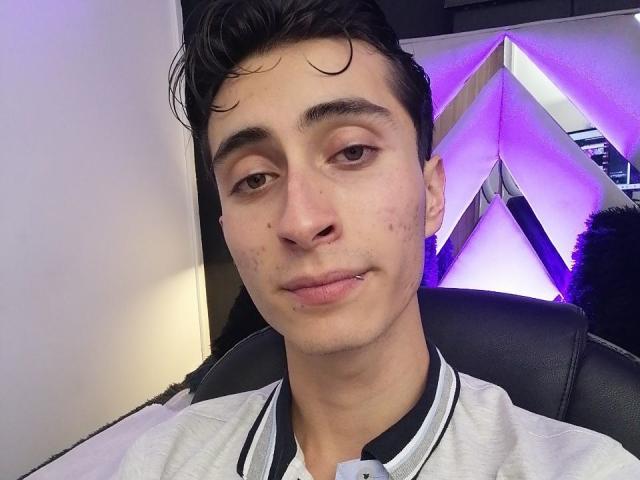 IthanSkin has a fair skin tone, dark wavy hair, brown eyes, small nose piercings, and is wearing a gray shirt with black trim. His face shows some acne.