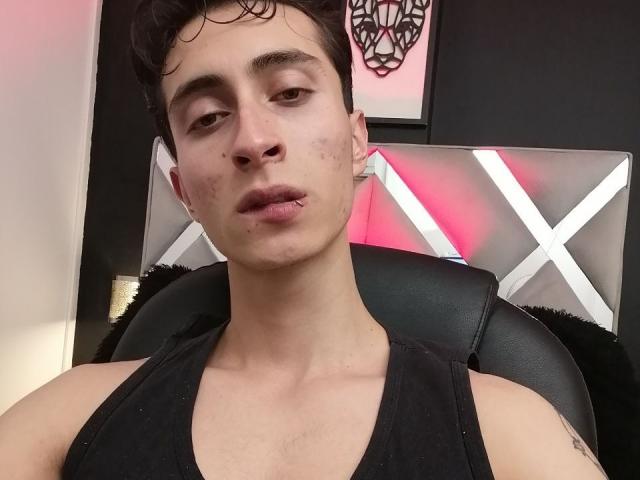IthanSkin tiene unos labios carnosos, una camiseta negra, cabello corto y oscuro, y un rostro ligeramente sucio con pequeñas manchas de acné. Está sentado en una silla de cuero negro..