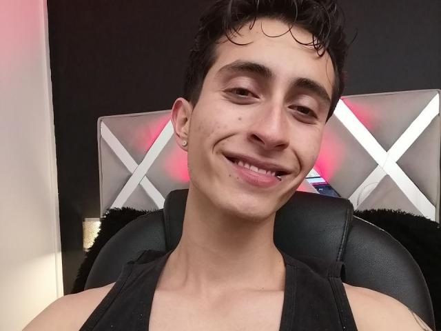 IthanSkin, een jonge man met krullend zwart haar, glimlacht met een klein gapje tussen zijn tanden, draagt een zwart tanktop en heeft een kleine oorbellen in zijn linker oor..