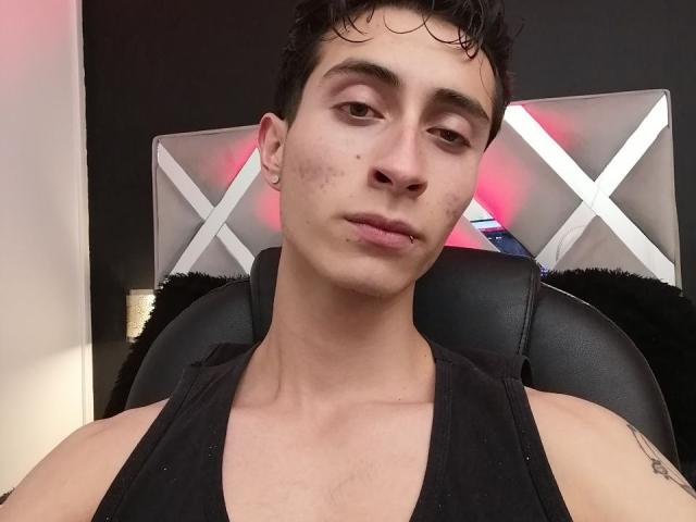 IthanSkin, a slim dude with short curly black hair, wearing a black tank top, has a small nose piercing, lip piercing, and a tattoo on his left arm.