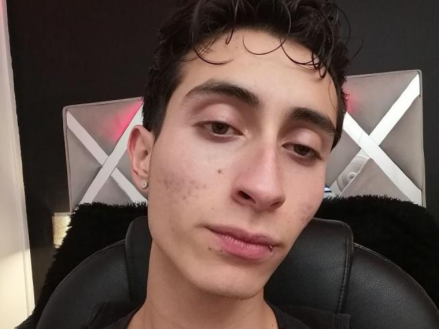 IthanSkin, con cabello negro rizado y corto, piel clara, pequeñas manchas de acné en la mejilla y un piercing en el labio, está sentado en una silla de cuero negro, luciendo ligeramente aburrido..