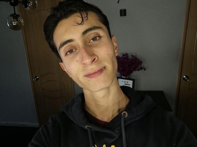 IthanSkin has short, dark hair, light skin, brown eyes, a small nose piercing, and a slight smirk. Wearing a black hoodie, with a wooden door in the background.