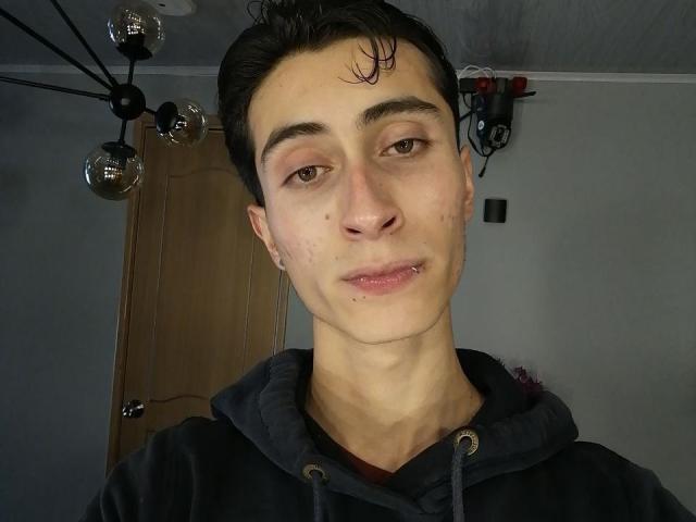 IthanSkin has a pale face with light brown eyes, short black hair, a small nose piercing, and a slight smile. Wearing a black hoodie, with acne and a cut on cheek.