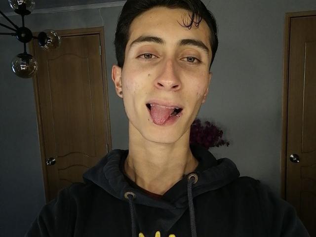 IthanSkin with short dark hair, sticking out his tongue, wearing a black hoodie, has a small bruise on his right cheek, and small earrings in a dimly lit room.