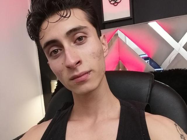 IthanSkin, mit kurzen, lockigen, schwarzen Haaren, einem Nasenpiercing und einem Lippenpiercing, trägt ein schwarzes Tanktop und sitzt in einem schwarzen Ledersessel, wobei er/sie in die Kamera schaut..