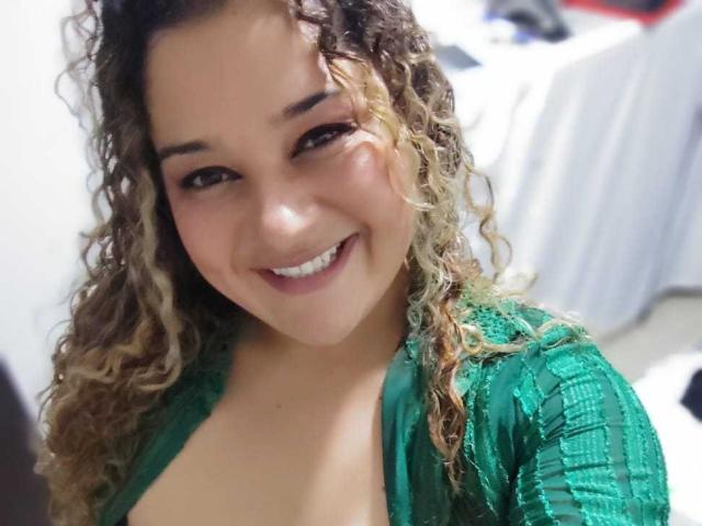 NataliaWendys with curly brown and blonde hair, smiling, wearing a green lace top, showing her tits, light skin, and brown eyes in a selfie.