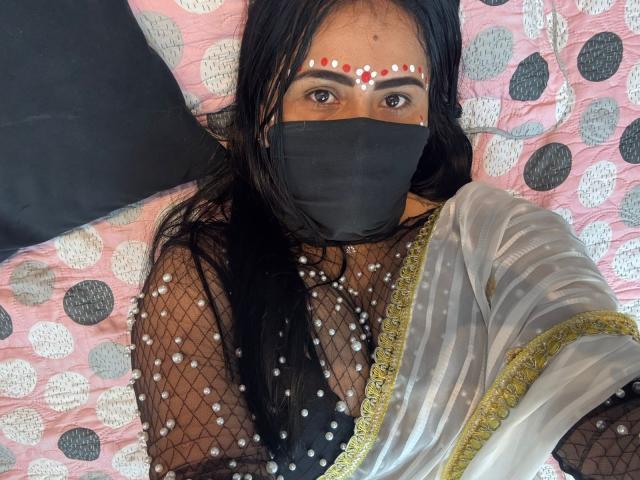 DaliaDubey with dark hair, brown eyes, and red dot makeup, wearing a black mask, see-through mesh top with pearl patterns, and a silver and gold saree, lying on a bed.