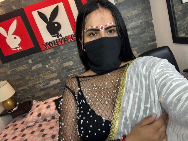 DaliaDubey with black mask, long black hair, face dotted with white pearls, wearing mesh top with white pearls, black bra, white striped cardigan, holding arm, dark background.