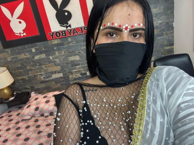 DaliaDubey with black mask, straight black hair, white and red dot forehead, fishnet top with white pearls, showing small tits, gray and white striped garment, stone wall background.