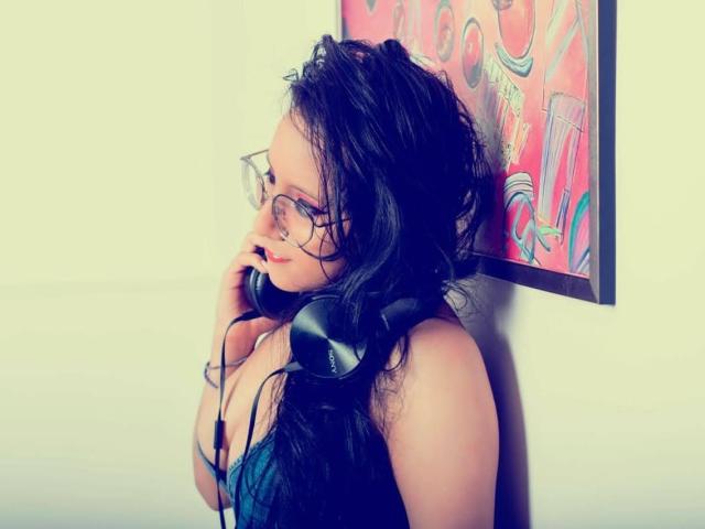 MiaKhalipso with wet, dark, curly hair wearing glasses and a blue tank top, holds black headphones to her ear. Her right hand touches her cheek. A colorful, abstract framed artwork is in the background. The image has a soft, pastel color palette with a slightly blurred, dreamy effect. MiaKhalipso's expression is relaxed and focused. The composition is centered, with MiaKhalipso slightly turned to the right.