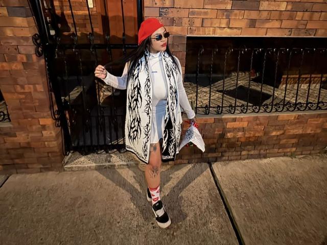 TessaMagic wears a red beanie, white turtleneck, black-and-white patterned coat, and white jeans. She's got striped socks and black-and-white sneakers, holding a newspaper.