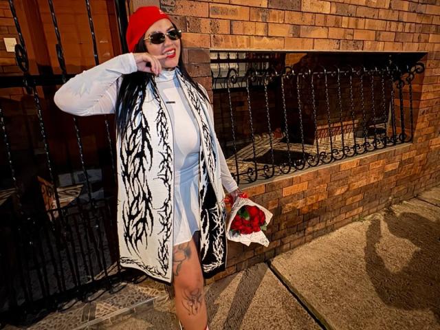 TessaMagic stands in front of a brick wall, wearing a red beanie, black-and-white patterned kimono, and white dress. She has sunglasses, long black hair, and a tattoo on her right thigh. She holds a bouquet of red roses. Her right arm is raised, touching her beanie. The brick wall has a black iron gate and a wooden door. The ground is textured and slightly shadowed. She's smiling, looking directly at the camera.