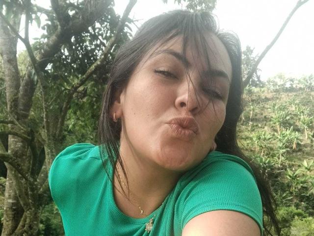 ScarlettOxford pouts her lips, making a silly face with her eyes closed, wearing a green shirt, her dark hair messy, standing outdoors with trees in the background.