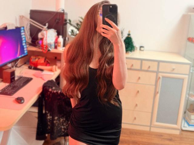 BiancaHotLove with long brown wavy hair, wearing a tight black dress, taking a mirror selfie with a phone, showing her back and ass, in a cluttered room.