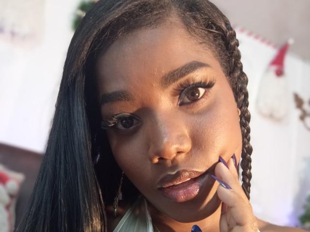 Caylin's face, dark skin, long black braids, thick eyebrows, big brown eyes with long lashes, glossy brown lips, blue nails, hand touching cheek, sunlight highlighting her features.
