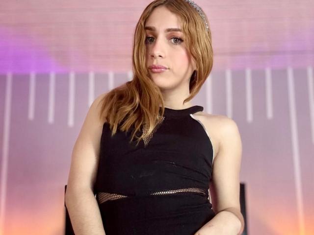 AndyDupont with light brown hair, wearing a black dress with mesh sides, looking serious, standing in front of a purple and pink-lit background. Andy's tits are modest.