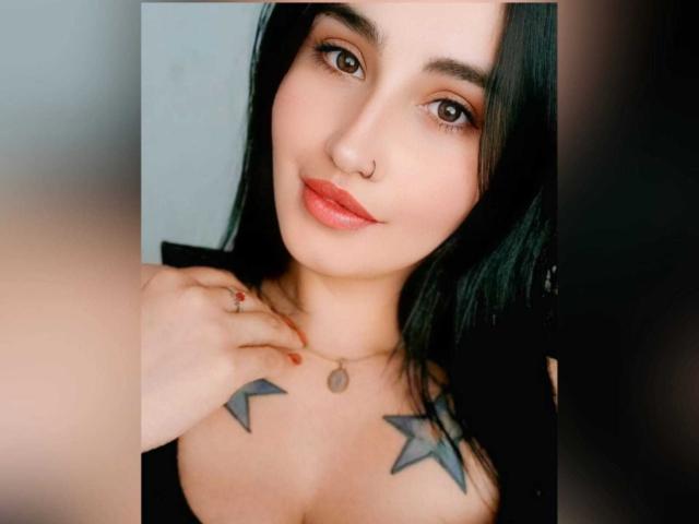 ScarletPriynce hat schwarzes Haar, braune Augen, einen Nasenring, roten Lippenstift und Tätowierungen an ihren Brüsten. Sie hält ihre Hand in der Nähe ihrer Brust und trägt ein schwarzes Oberteil..