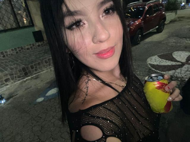 AdelleVelvet with long black hair, wearing a sheer black top, holding a yellow soda can, has a small nose ring and a tattoo on her right shoulder, smiling.