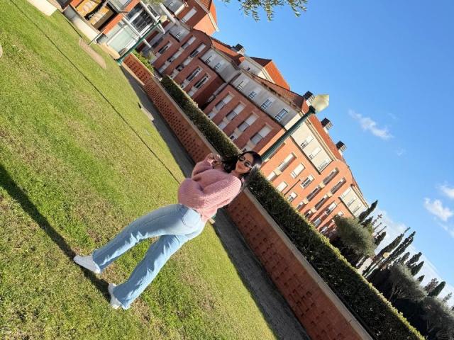 IsabellaJames stands on grass, arms crossed, wearing a pink sweater, blue jeans, and white sneakers. She has dark hair and a slight smile, with a red brick wall behind.