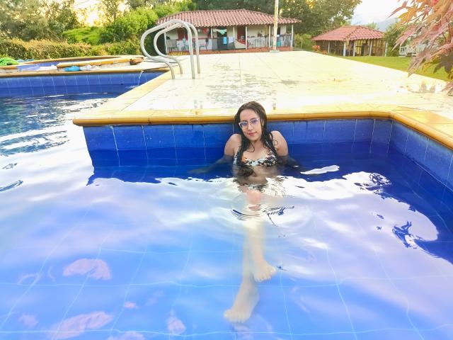 MiaKhalipso is in a blue-tiled pool, half-submerged with only her upper body visible. She wears a black and white patterned bikini top and glasses, with dark hair and light skin. Her legs are extended in the water. The pool has a wooden edge and a metal ladder. The background includes a red-tiled roof building and greenery. Bright sunlight reflects on the water. The image is clear and detailed, emphasizing the blue tiles and the reflective water surface.