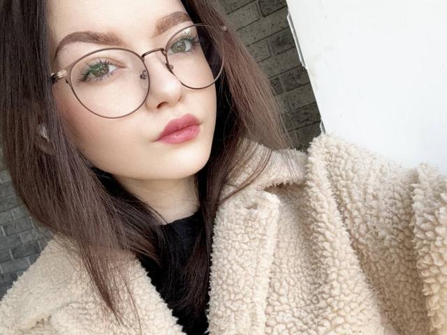 WeksaMur a les yeux vert clair, porte des lunettes marron, a les lèvres roses et est vêtue d'un manteau beige duveteux. Elle a les cheveux brun foncé et porte un haut noir. Son visage est légèrement incliné..