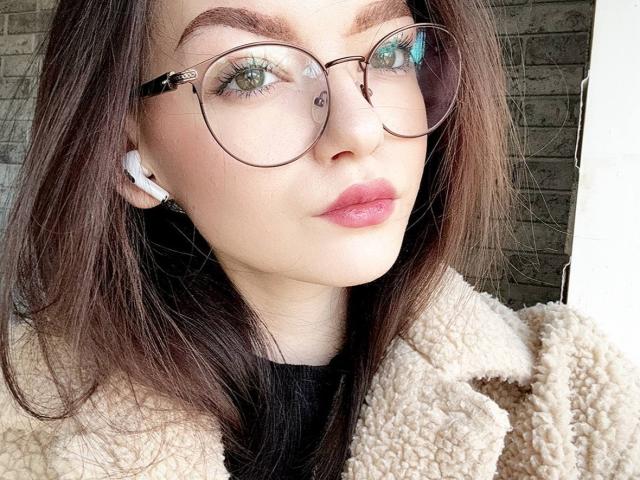 WeksaMur with round glasses, white earbuds, pink lips, and brown hair, wearing a fluffy beige coat, has green eyes and a cute, relaxed expression.